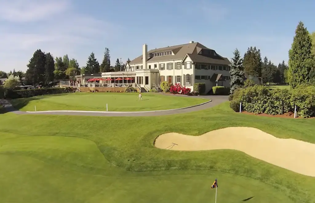 Wide view of a golf clubhouse overlooking a putting green, practice bunker, and manicured fairway on a sunny day