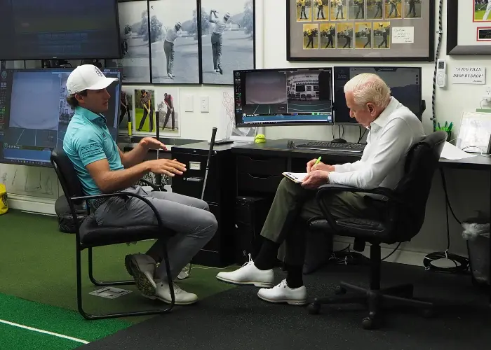 Indoor golf coaching session where an instructor reviews swing footage on monitors while discussing technique and taking notes