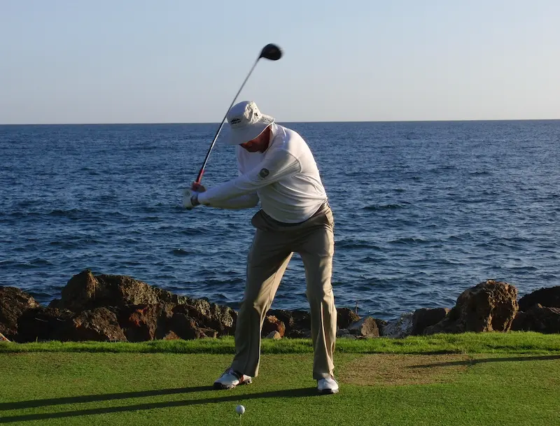 Golfer demonstrating a full swing on a practice tee with the ocean and rocky shoreline in the background