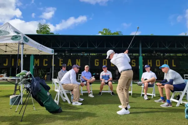 Golf instructor demonstrating a swing technique to a small group of students seated on a practice green during an in-person training session