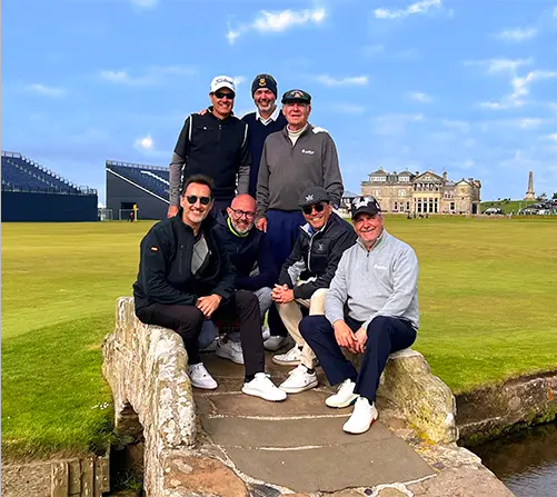 Group of golfers seated and standing together on a stone bridge at a historic links-style golf course, captured during a relaxed group visit in a scenic outdoor setting