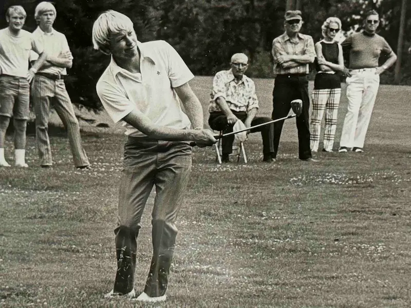 Vintage black and white photograph showing a golfer striking a shot on a grass course while spectators watch in the background