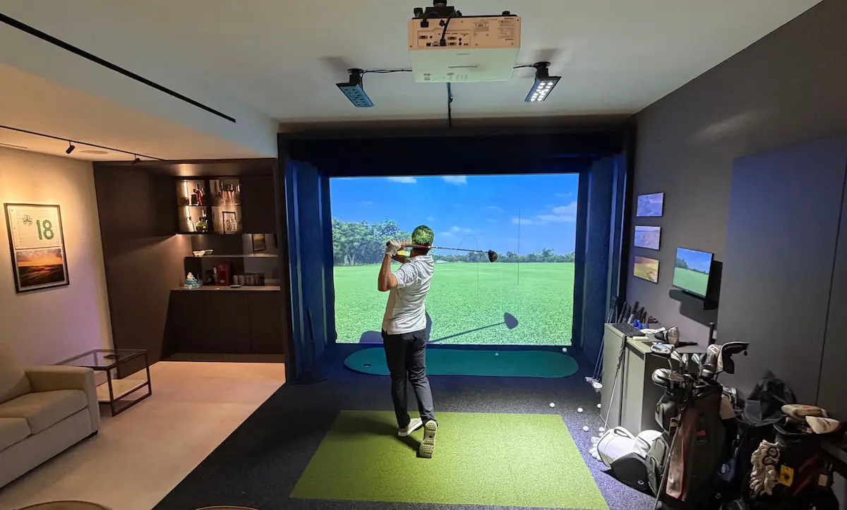 Golfer completing a swing inside an indoor simulator room, with a projected course display and practice equipment visible in a modern training environment