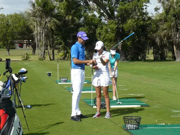Golf instructor reviewing swing technique with a student on the driving range while another golfer practices in the background