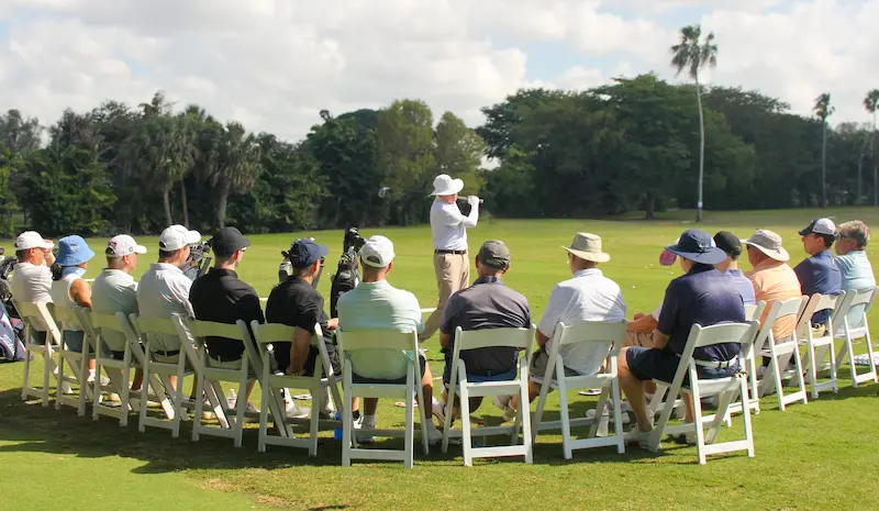 Golf instructor demonstrating a swing to a seated group of students during an outdoor training session on a practice range